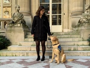 Justine est à côté de Snow qui est assise avec son body élève chien guide. Les deux se regardent, Justine sourit.